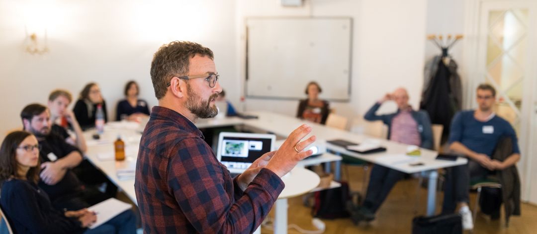 In einem Seminarraum sitzen viele Personen an einem U-Förmig aufgestellten Tisch. Sieh hören einer weiteren Person zu, die einen Vortrag hält.