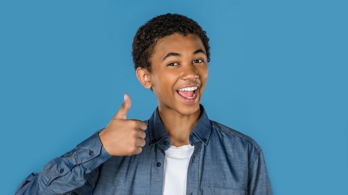 Ein jugendlicher steht vor einem blauen Hintergrund, lacht fröhlich und zeigt "Daumen hoch".