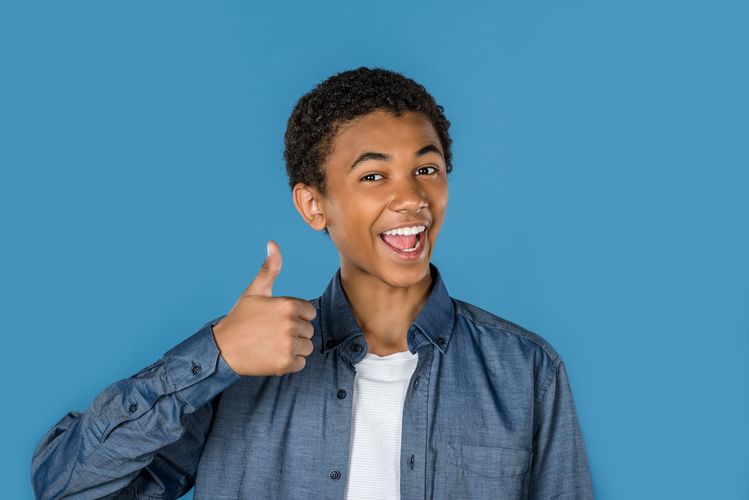 Ein jugendlicher steht vor einem blauen Hintergrund, lacht fröhlich und zeigt "Daumen hoch".