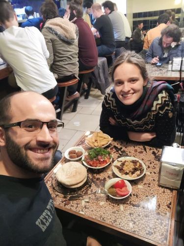 Ein Mann und eine Frau sitzen sich am Tisch gegenüber. Der Mann hält das Handy in die Höhe, um ein Selfie der beiden zu schießen. Auf dem Tisch zwischen ihnen steht Essen in diversen kleinen Schalen.