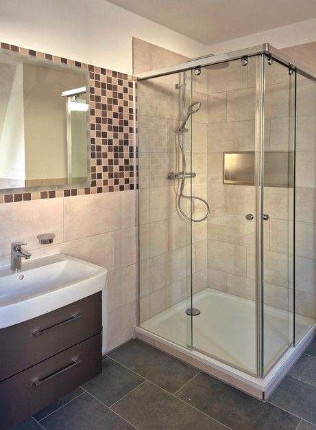 enlarge the image: Modern bathroom featuring a corner glass shower with a handheld showerhead, a wall-mounted sink with a vanity unit, a large mirror, and tiled flooring along with a wall showcasing decorative accents. Photo: Thomas Biermann