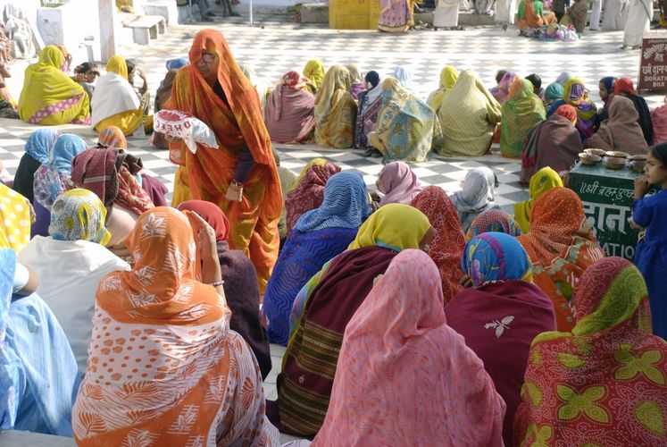 Viele Frauen sitzen in bunten Saris zusammen. Sie sind von Hinten fotografiert. Eine alte Frau geht zwischen ihnen hindurch mit einer Tüte in der Hand.
