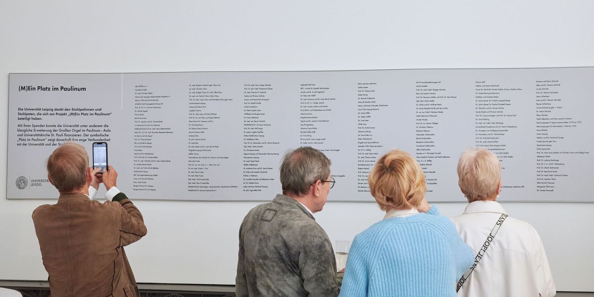 zur Vergrößerungsansicht des Bildes: zwei Stuhlpaten und zwei Stuhlpatinnen stehen vor der Stuhlpatentafel im Paulinum und lesen die Namen, die auf der Tafel veröffentlicht sind, ein Mann fotografiert