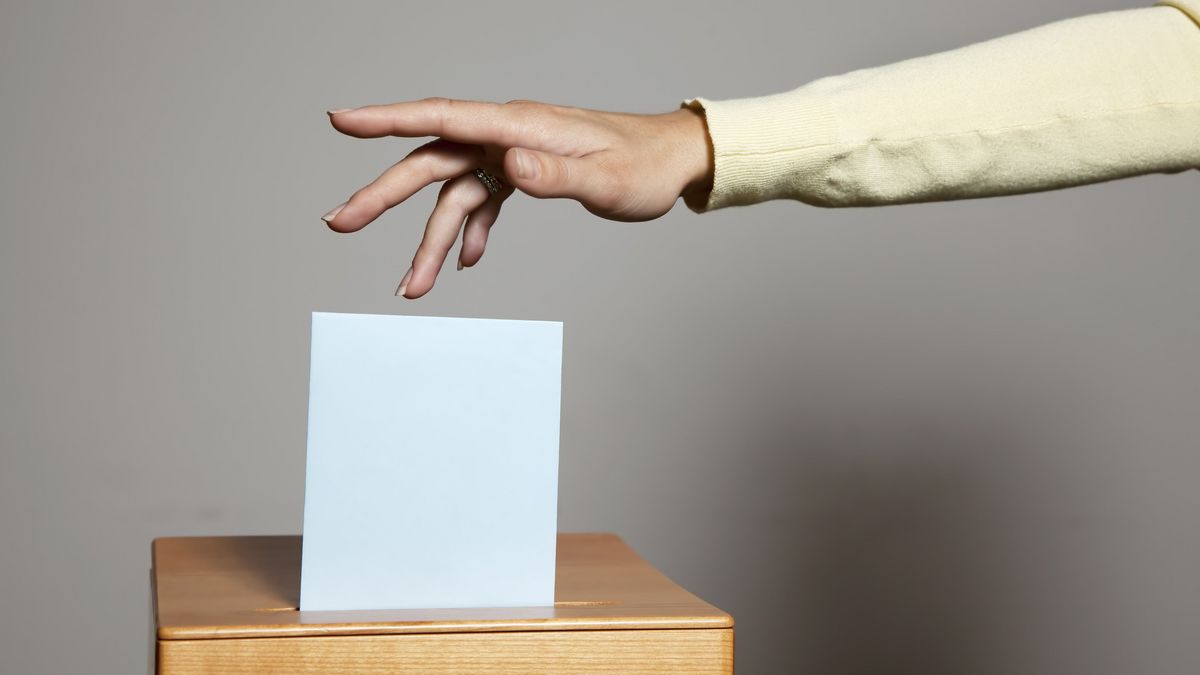 Alternativtext: Ansicht einer Wahlurne in die eine Stimmabgabe erfolgt, eine weiblich gelesen Hand wirft einen Briefumschlag ein.