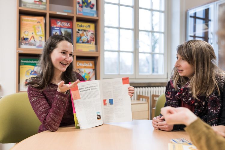 Eine Studentin sitzt an einem Tisch und erklärt etwas zu einem Buch mit der aufgeschlagenen Seite "Lesen und schreiben in heterogenen Gruppen". Sie hält es hoch, damit es ihre Kommilitonen und Kommilitoninnen am Tisch sehen können, Foto: Christian Hüller 