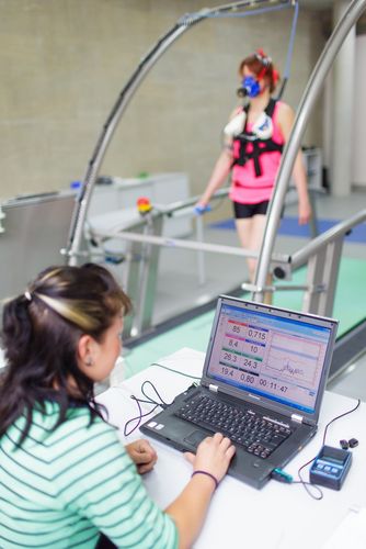 Studentin steht vor einem Laptop und im Hintergrund sieht man eine verkabelte Probantin auf einem Laufband, Foto: Christian Hüller