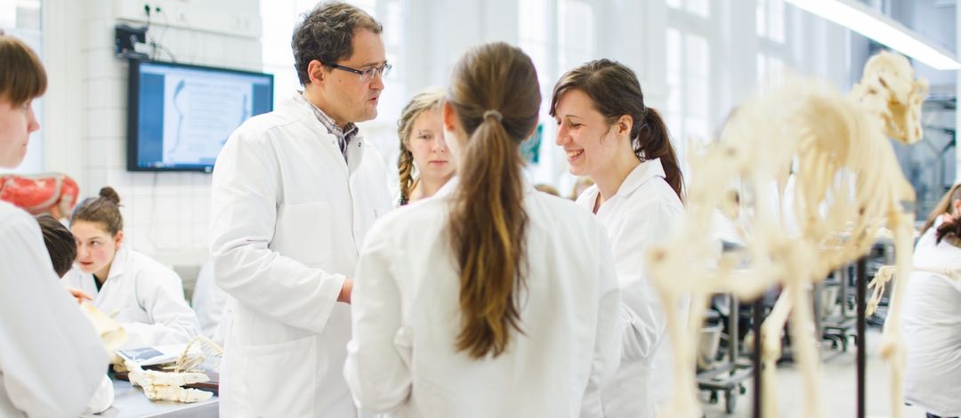 Foto: Studierende in Arztkittel gekleidet stehen im Anatomiesaal und unterhalten sich