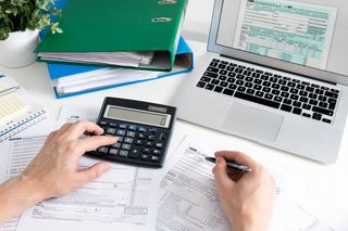 Farbfoto: Blick auf einen Schreibtisch mit Laptop, zwei Aktenordnern, Taschenrechner, Formlare und zwei Hände, die rechte Hand hält einen Stift