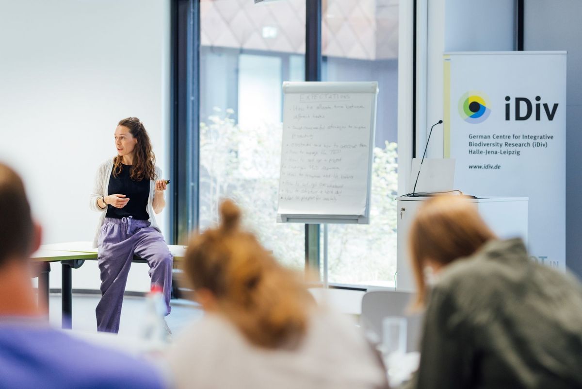 enlarge the image: Farbfoto: Eine Frau sitzt lehnt an einem Tisch und spricht zu Studierenden. Im Hintergrund steht ein Banner mit der Aufschrift "iDiv".