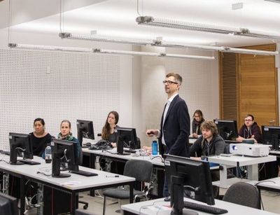 Abgebildet ist ein heller Raum mit vielen Computerarbeitsplätzen an denen Studierende sitzen und dem Dozenten zuhören.
