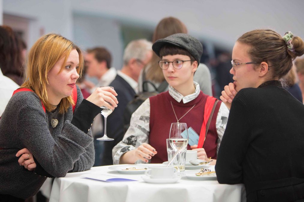 zur Vergrößerungsansicht des Bildes: drei Stipendiatinnen stehen am Stehtisch, unterhalten sich, auf dem Tisch stehen Gläser und Geschirr