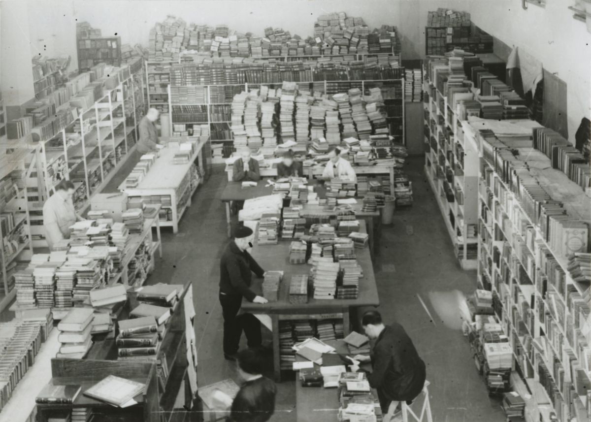 zur Vergrößerungsansicht des Bildes: historische schwarz-weiss-Aufnahme: viele gestapelte Bücher und einige Menschen darin blätternd