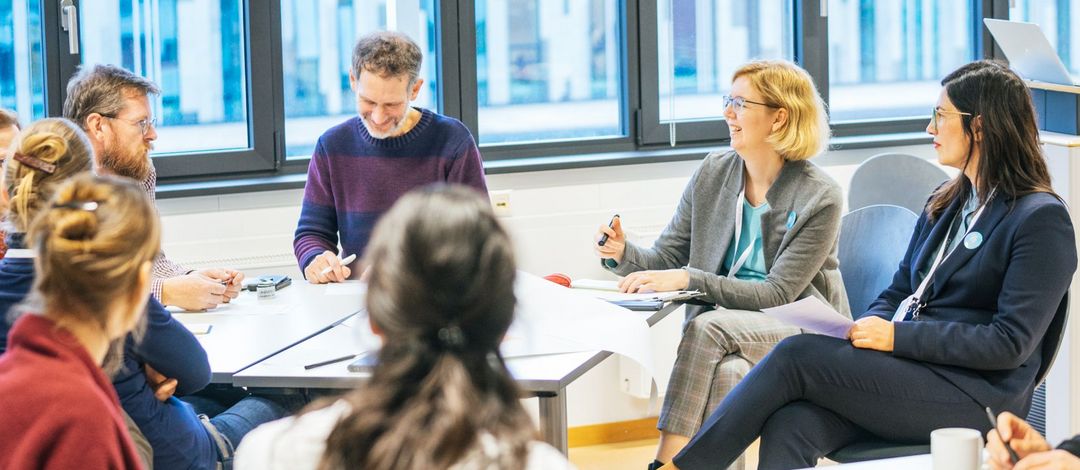 Farbfoto: Acht Personen unterschiedlichen Geschlechts sitzen in einer Runde am Tisch und diskutieren miteinander.