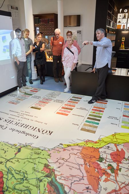 Farbfoto: Kustos Frank Bach gibt den Gästen Informationen zur Sammlung. Die Personen stehen im Ausstellungsraum, auf dem Fußboden ist eine große Landkarte zu sehen.