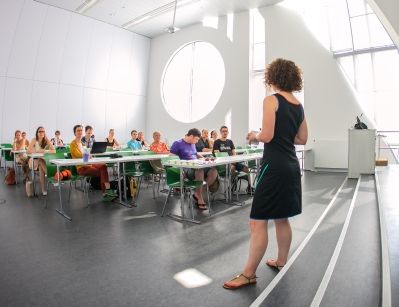 Foto: eine Vortragende steht in Front von Zuhörenden an Tischen sitzend