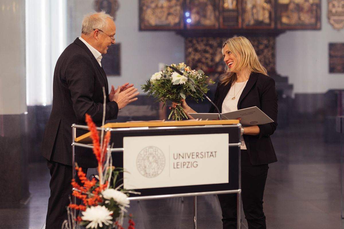 Prof. Dr. Thomas Lenk steht rechts im Bild. Prof. Dr. Elisa Marie Hoven steht links im Bild. Er überreicht ihr einen Blumenstrauß und gratuliert ihr.