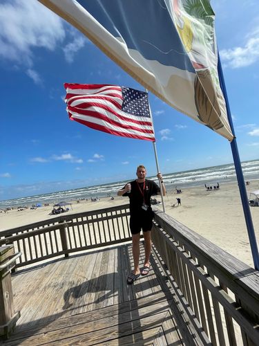 Der Student Jonas steht mit einem Getränk in einer Dose in der linken Hand und einer USA-Flagge in der rechten Hand auf einem Steg, der über einen Strand ragt. Er lächelt in die Kamera. Im Hintergrund sind Strand und Meer zu sehen.