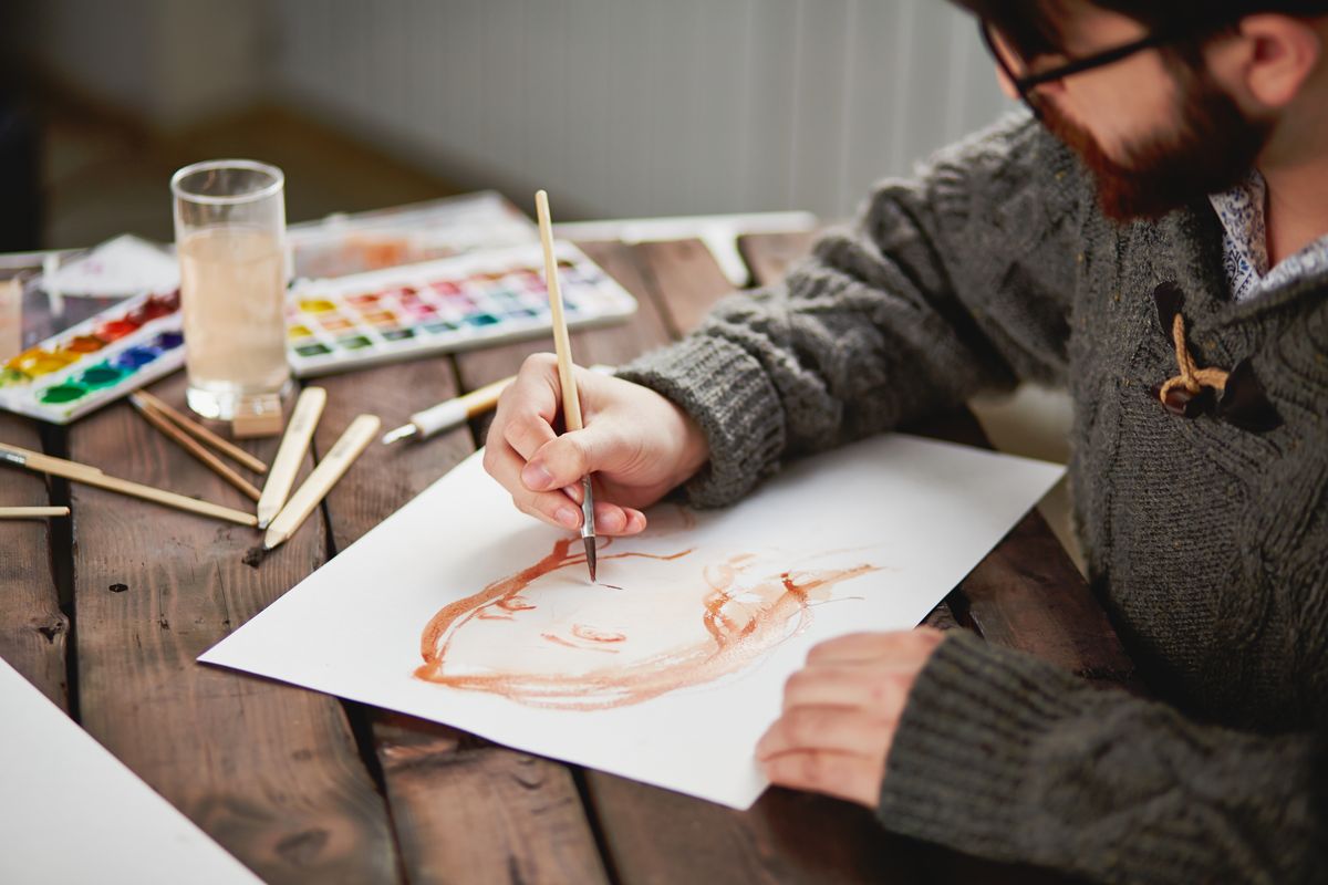 Zu sehen ist ein junger Mann mit Bart, der an einem Tisch sitzt und mit Wasserfarben auf Papier malt.