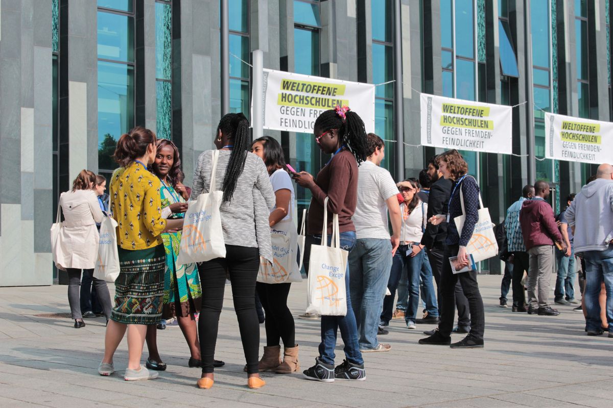 Zu sehen auf dem Foto ist eine internationale Menschengruppe auf dem Augustusplatz, im Hintergrund ist das Universitätsgebäude sichtbar. Die Personen scheinen im regen Austausch zu sein, im Hintergrund prangen Plakate mit der Aufschrift "Weltoffene Hochschulen gegen Fremdenfeindlichkeit".