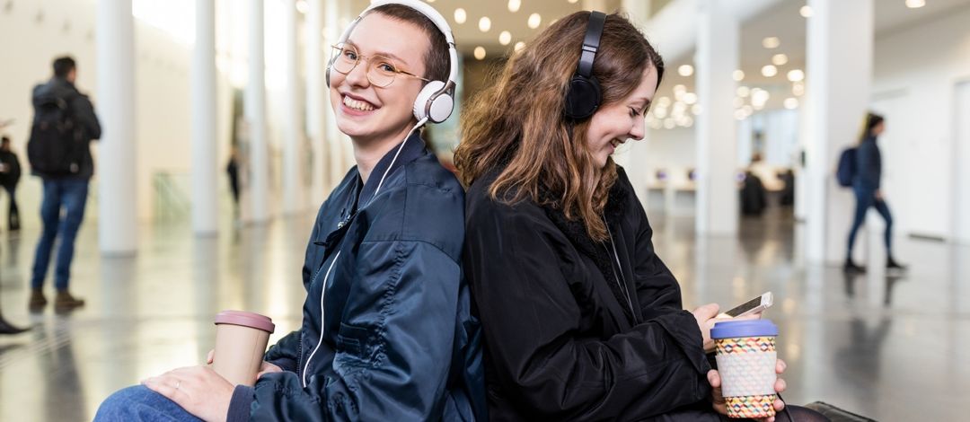 Zwei Studentinen sitzen Rücken an Rücken im Augusteum und haben Kopfhörer auf. Die linke Studentin blickt lachend in die Kamera. Die rechte Studentin schaut lächenlnd auf ihr Smartphone, welches sie in der Hand hält.