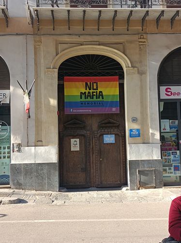 Eingang mit hölzernen Tür, daran hängt eine Regenbogen-Fahne mit der Beschriftung 'No Mafia Memorial', bei Tag