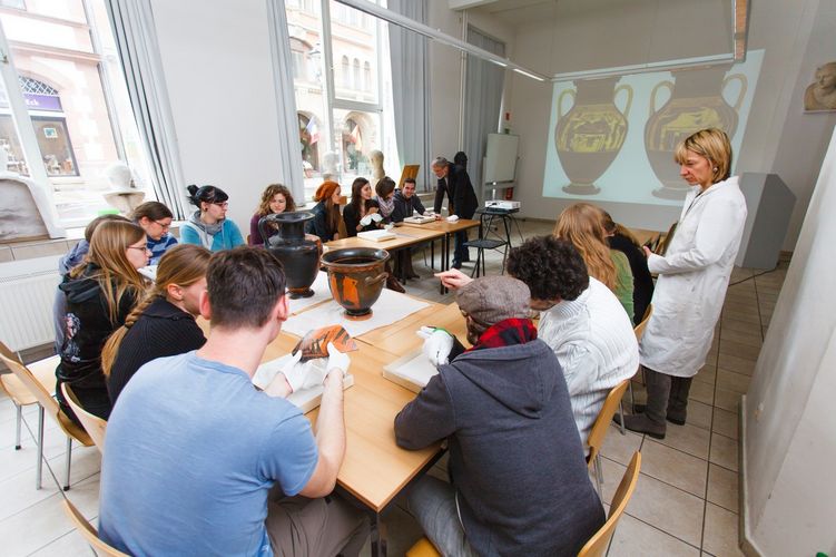 In der Mitte des Seminarraums stehen antike Vasen auf Tischen und Studierende sitzen darum und schauen sich mit Handschuhe Scherben an, Foto: Christian Hüller