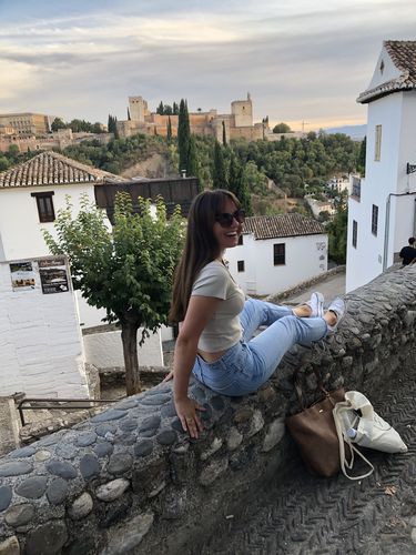 Eine lachende Frau sitzt auf einer niedrigen Steinmauer. Sie schaut in die Kamera. Im Hintergrund ist eine Stadt zu sehen. 