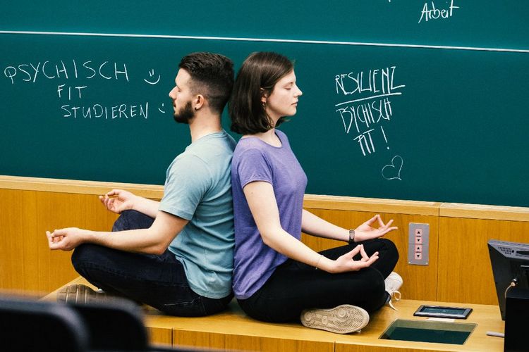 Farbfoto: Eine Frau und ein Mann meditieren in einem Hörsaal vor einer Tafel. Sie sitzen dabei im Schneidersitz auf dem Pult des Dozierenden. Sie haben die Augen geschlossen und sitzen Rücken an Rücken.uf einem Tisch