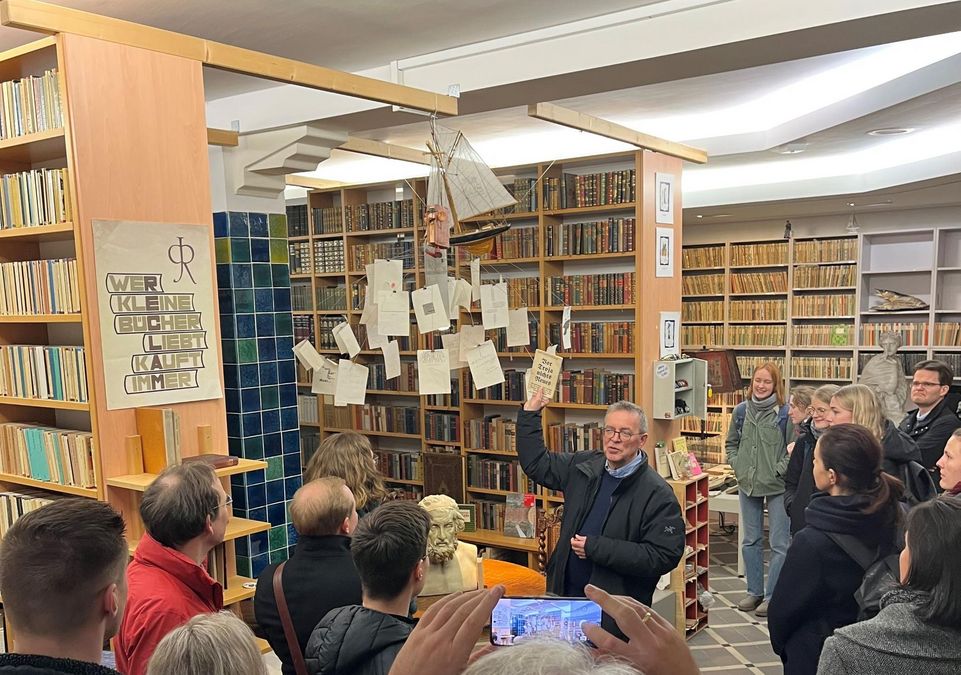 zur Vergrößerungsansicht des Bildes: Förderer und Stipendiaten stehen im Bibliotop und hören den Ausführungen von Professor Lokatis, der in der Mitte zu steht, zu, im Hintergrund sind Bücherregale zu sehen