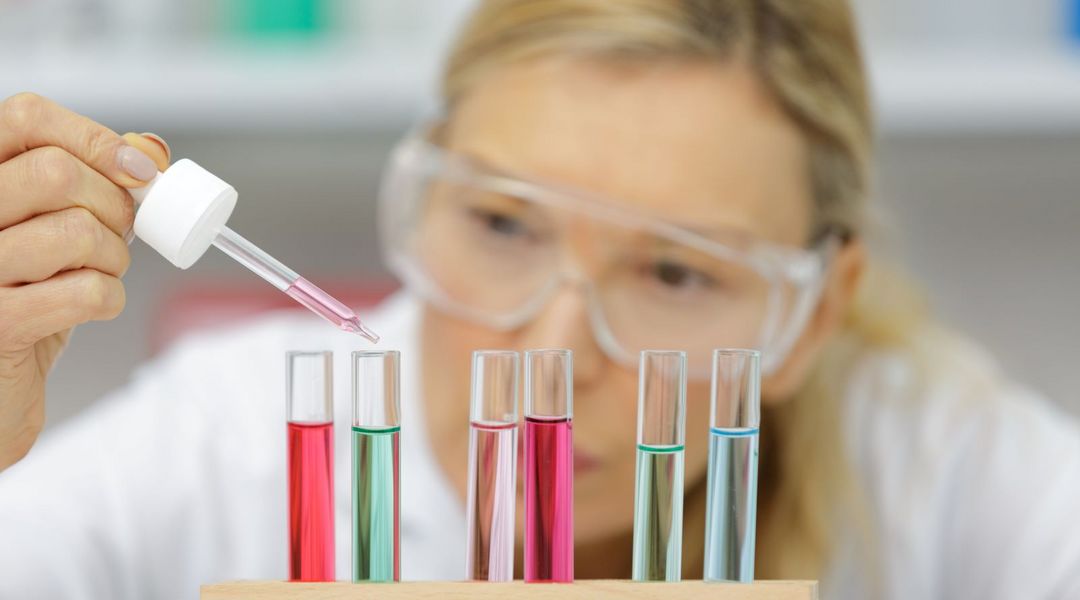 Farbfoto: Eine Frau mit weißem Laborkittel und Schutzbrille steht hinter mehreren Reagenzgläsern. In der Hand hält sie eine Pipette und gibt rote Flüssigkeit in eines der Reagenzgläser.