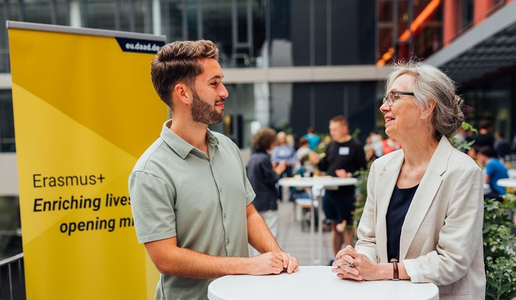 Eine Frau und ein Mann stehen gemeinsam an einem Stehtisch und blicken sich an. Hinter Ihnen sieht man ein Roll-Up des Förderprogramms Erasmus+. Weiter im Hintergrund erkennt man Silhouetten von Menschen, die an einer Veranstaltung teilnehmen. 