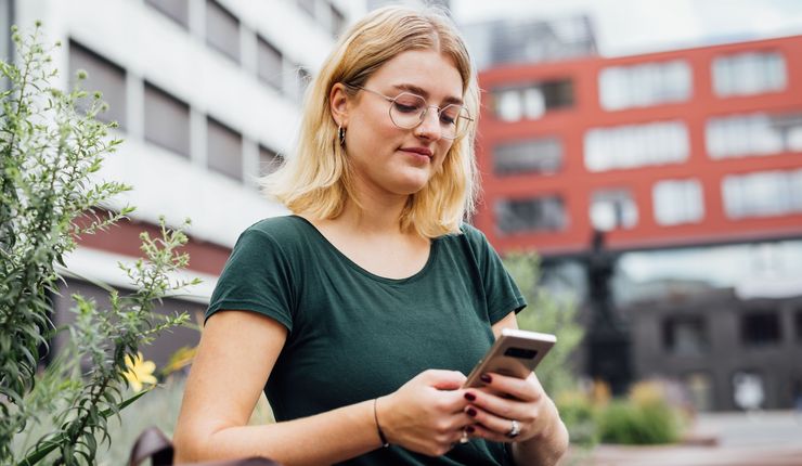 Studentin sitzt auf auf einer Bank auf dem Campus und schaut auf ihr Handy, Foto: Christian Hüller