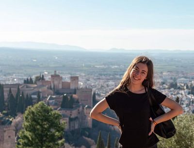 Farbfoto: Eine Frau lächelt in die Kamera. Im Hintergrund befindet sich ein burgenähnliches Gebäude und eine Landschaft mit Häusern