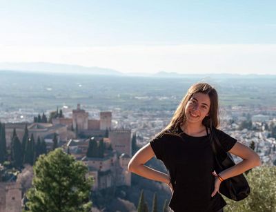 Farbfoto: Eine Frau lächelt in die Kamera. Im Hintergrund befindet sich ein burgenähnliches Gebäude und eine Landschaft mit Häusern