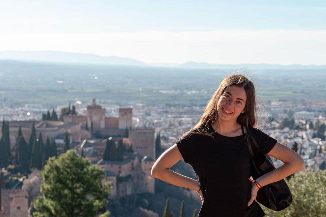 zur Vergrößerungsansicht des Bildes: Farbfoto: Eine Frau lächelt in die Kamera. Im Hintergrund befindet sich ein burgenähnliches Gebäude und eine Landschaft mit Häusern