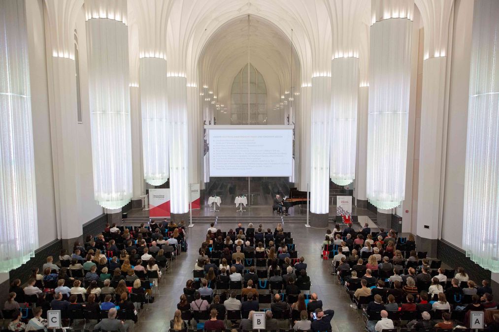 zur Vergrößerungsansicht des Bildes: Förderer und Stipendiaten sitzen in Reihen und Blicken Richtung Bühne, dort steht ein Flügel auf dem Claudius Bruns spielt, die Aula ist hell erleuchtet