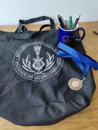 Ein schwarzer Jutebeutel mit einem silbernen Schullogo, eine Medaille, eine blaue Tasse mit Schullogo stehen auf einem Tisch. In der Tasse befinden sich vier Kugelschreiber.