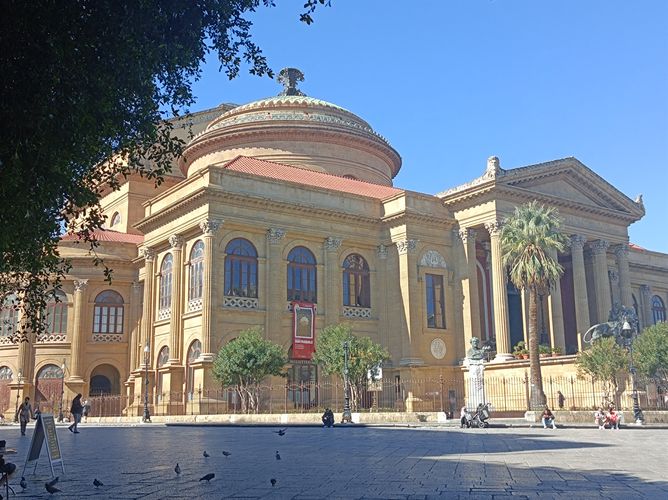 Sandsteinfarbenes Theater mit Kuppel und Säulen, davor ein Platz, Bei Sonnenschein