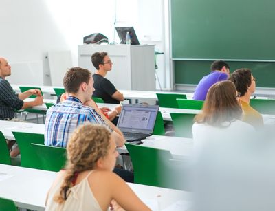 Studierende sitzen im Hörsaal an Tischen verteilt und folgen einer Vorlesung