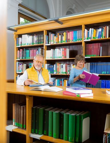 Farbfoto: Blick in eine Bibliothek. Ein älterer Mann mit gelber Weste und weißem Hemd sowie eine Frau in einem blauen Kleid stehen lesend vor einem Bücherregal aus hellem Holz