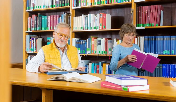 Farbfoto: Blick in eine Bibliothek. Ein älterer Mann mit gelber Weste und weißem Hemd sowie eine Frau in einem blauen Kleid stehen lesend vor einem Bücherregal aus hellem Holz