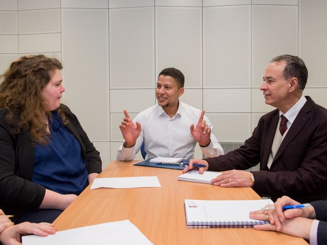 Ein Mann sitzt zwischen zwei Gesprächspartnern, hält die Hände mit den Zeigefingern nach oben und blickt eine Gesprächspartnerin an. Foto: Bodo Tiedemann