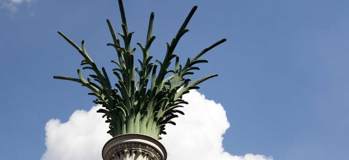 Detailaufnahme: Palmenwedel auf einer Säule vor blauem Himmel mit weißer Wolke im Hintergrund