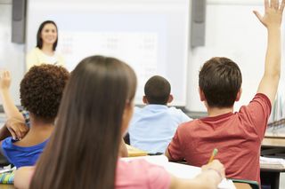 Farbfotografie aus dem hinteren Bereich eines Klassenzimmers mit Kindern und einer Lehrerin, die an einer Tafel steht.