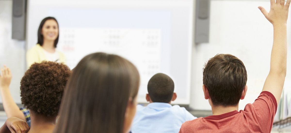 Farbfotografie aus dem hinteren Bereich eines Klassenzimmers mit Kindern und einer Lehrerin, die an einer Tafel steht.