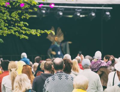 Man sieht Menschen, die sich in einer großen Gruppe bei einem Fest versammeln. Im Hintergrund sieht man eine Bühne mit Künstlern.