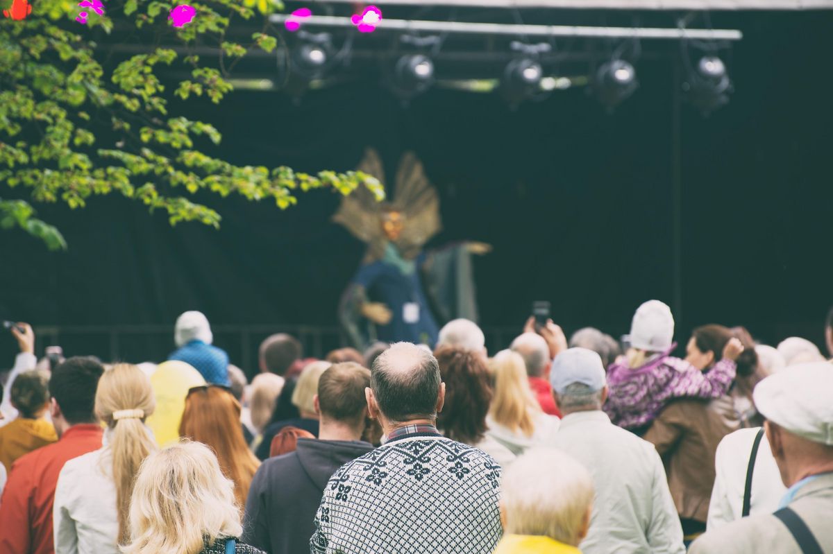zur Vergrößerungsansicht des Bildes: Man sieht Menschen, die sich in einer großen Gruppe bei einem Fest versammeln. Im Hintergrund sieht man eine Bühne mit Künstlern.
