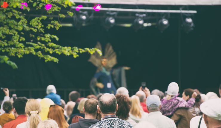 Man sieht Menschen, die sich in einer großen Gruppe bei einem Fest versammeln. Im Hintergrund sieht man eine Bühne mit Künstlern.