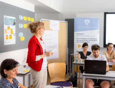 Farbfoto: eine Frau steht seitlich vor einer Tafel und spricht zum Publikum. 