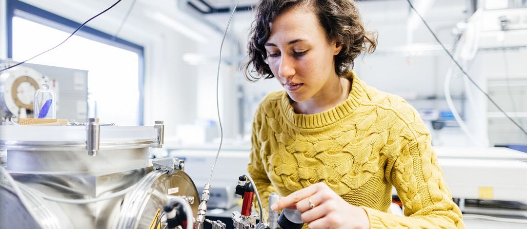 Farbfoto: Eine Frau steht in einem Labor vor einem technischen Gerät und stellt dort etwas ein.